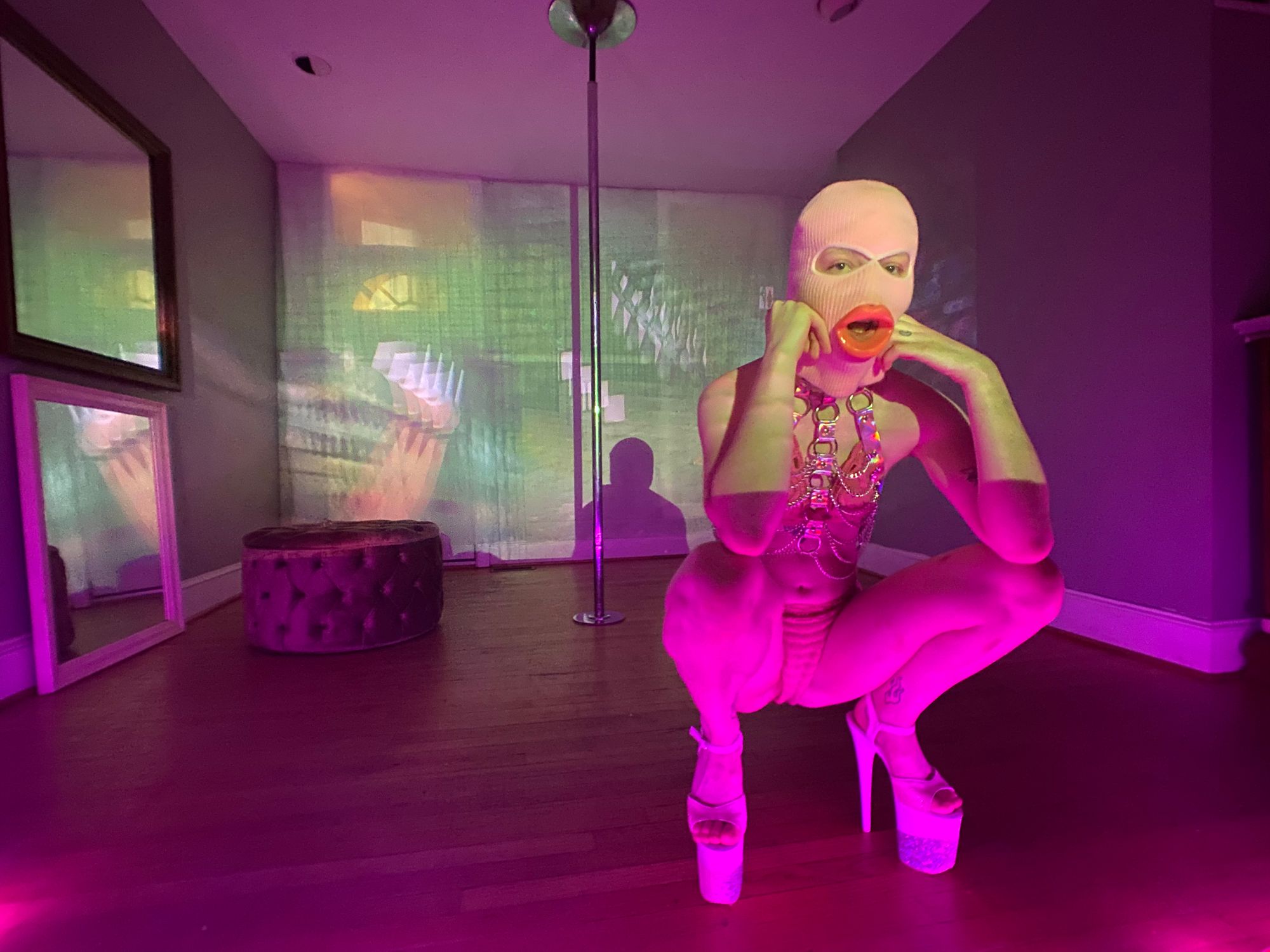 Squiggles is crouching in front of a mostly bare room. There is a pole, and ottoman, and a video of IMVU gameplay projected across the wall.