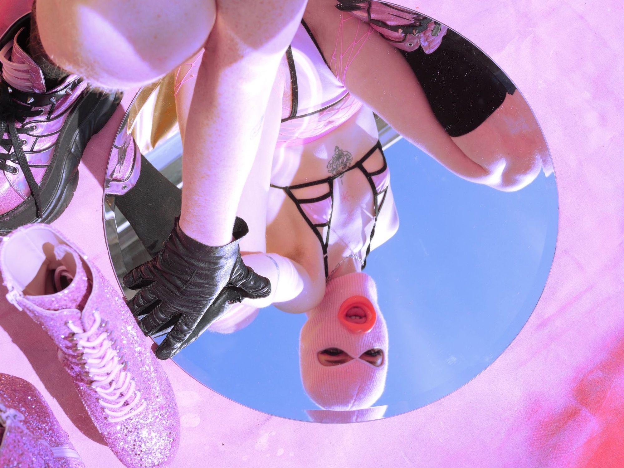 Squiggles is crouched above a round mirror, which reflects their masked face and the bright blue sky back to them. The background is pink, with pink glitter platform heels just in frame.