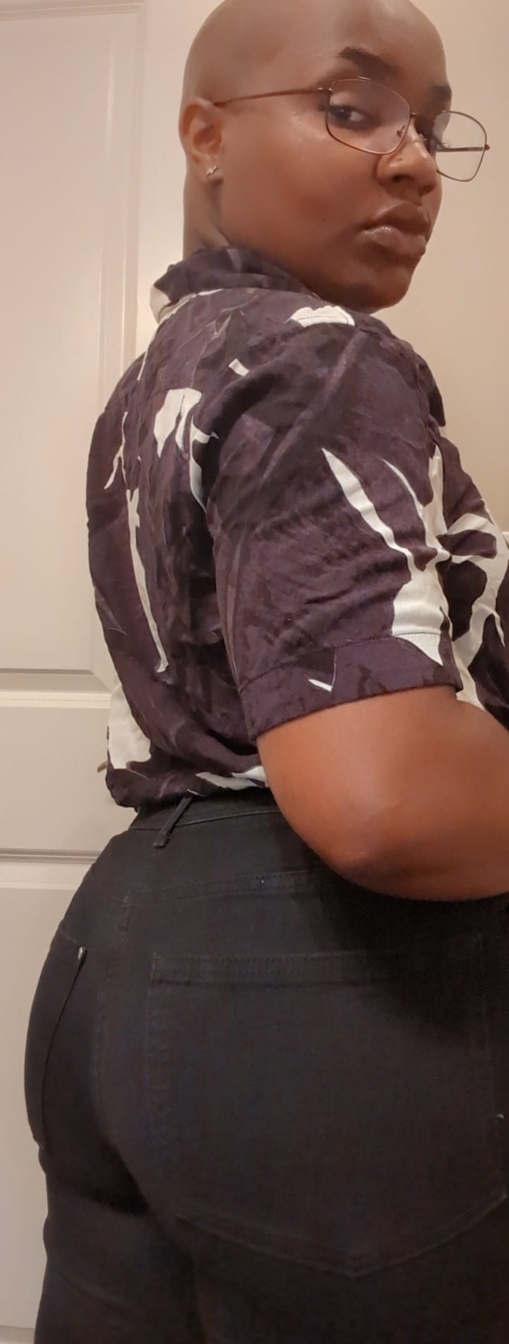 Devon Rose, a Black, trans person looking back to the camera in a patterned shirt and black jeans.