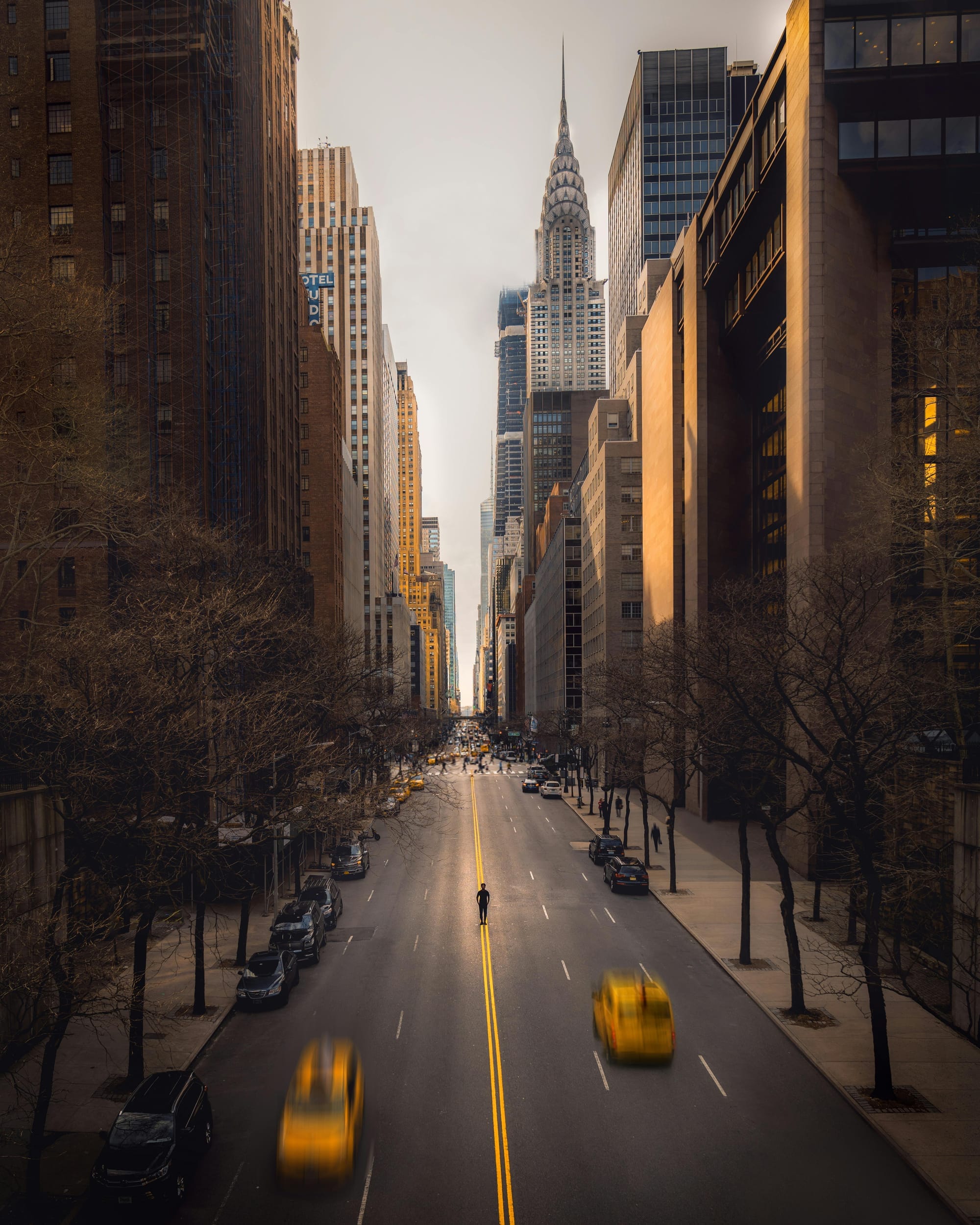 A New York City street with a figure standing in the middle of the road and two cabs passing them.