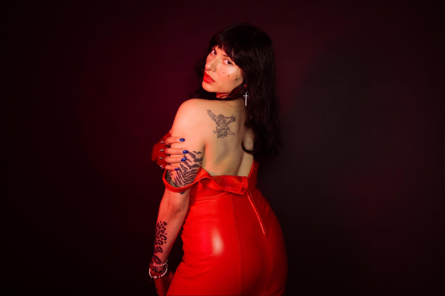 Dominatrix Nomi Fatale looks over her shoulder. She is wearing a red leather dress with one strap falling down.