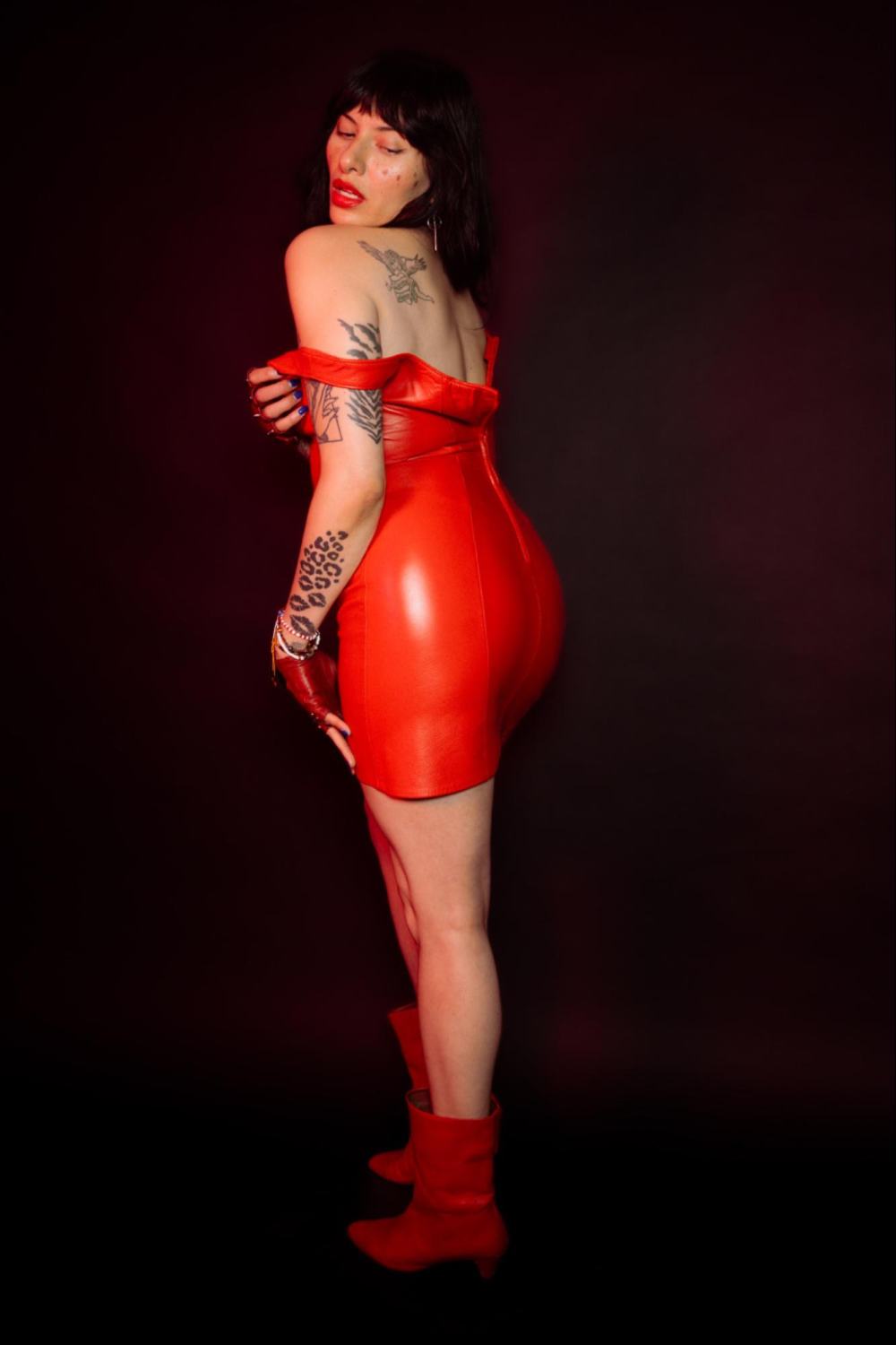 Image of Dominatrix Nomi Fatale looking over her shoulder. She is wearing a red leather dress and matching boots.