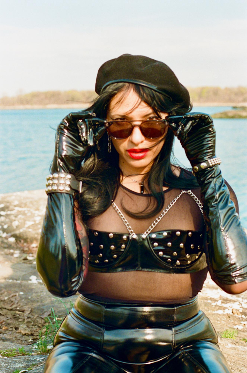 Photo of Nomi Fatale sitting on rocks by the ocean, dressed in shiny black skirt, bra, and gloves. She is wearing a beret and sunglasses.