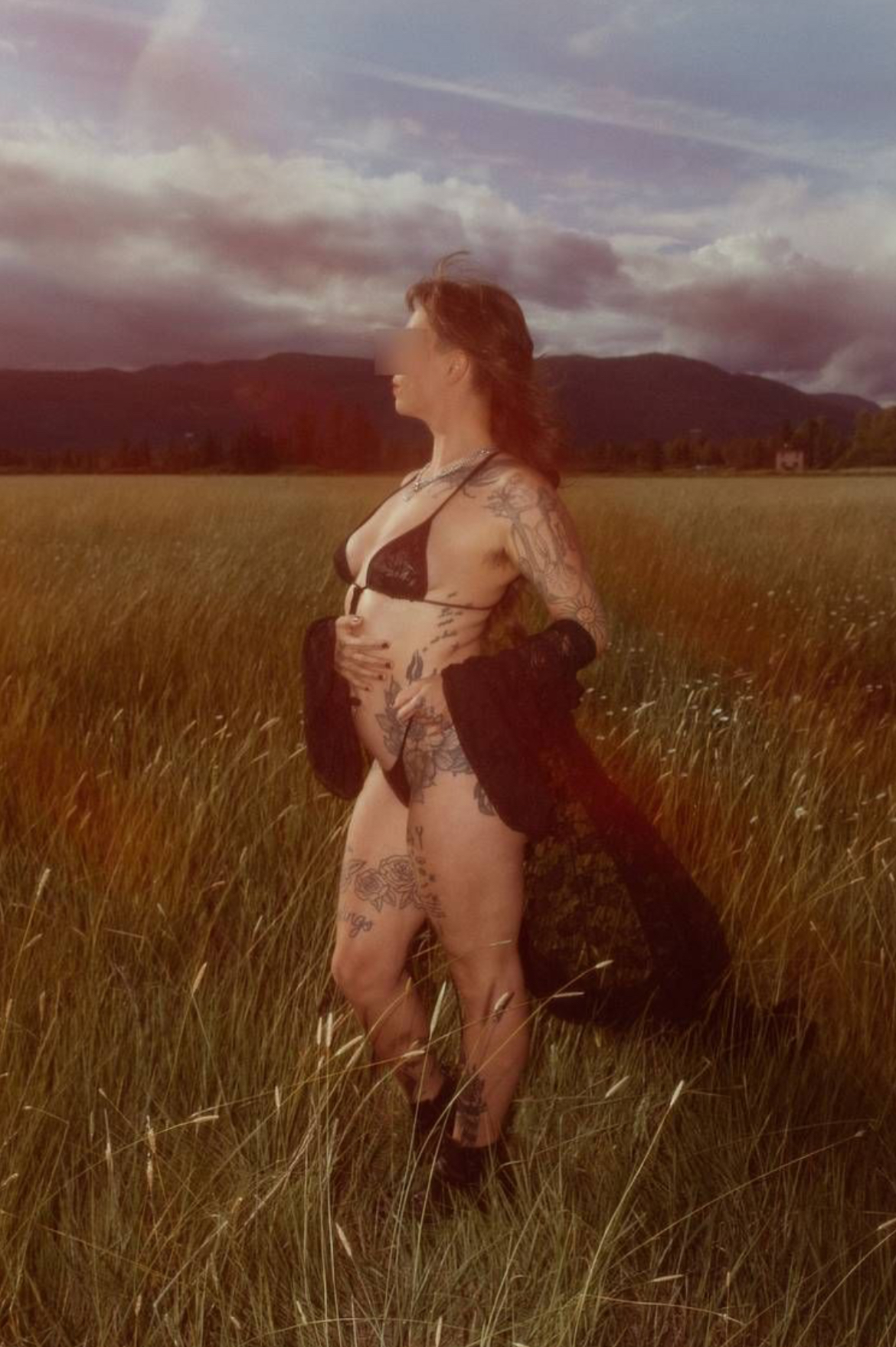 Rae De Rosa stands in a field of long grass, wearing black lingerie and a black robe.
