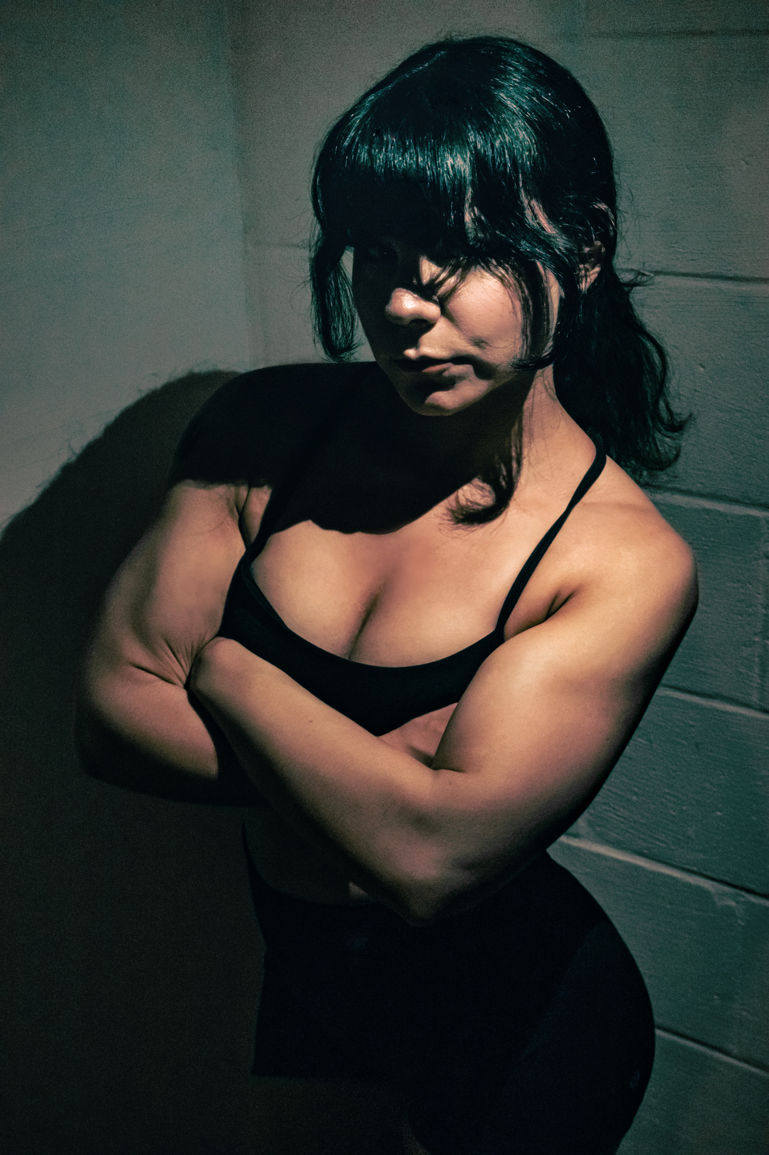 Mistress Blaze stands with her arms crossed, her muscular biceps emphasised, wearing a simple black dress.
