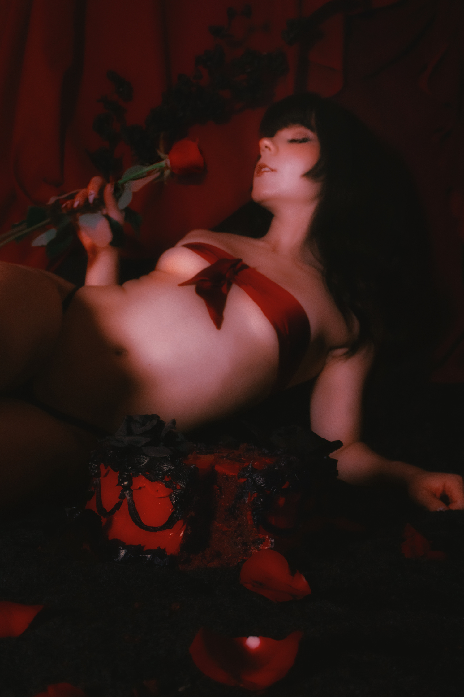 Mistress Blaze reclines, a ribbon tied around her breasts, holding a red rose.