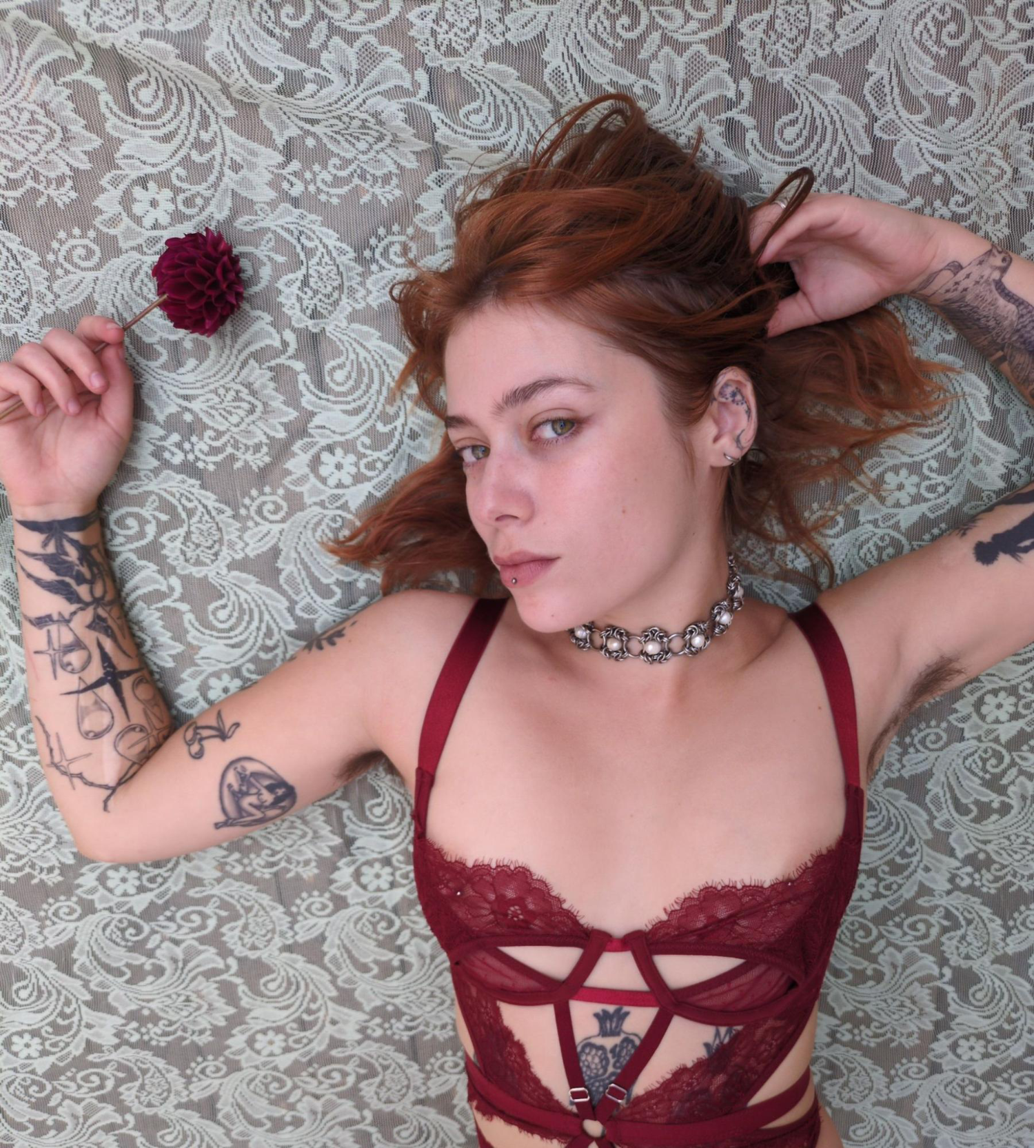 Seattle Escort Chiara Novo lays on a lace blanket, holding a rose and dressed in red lingerie.