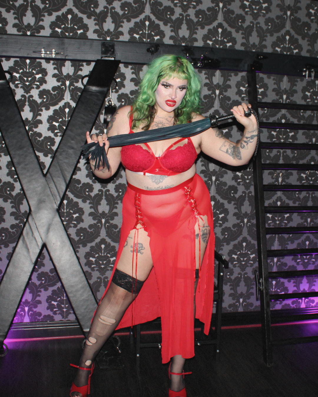 Mistress Ritual holds a leather flogger while standing in front of a St Andrews cross.